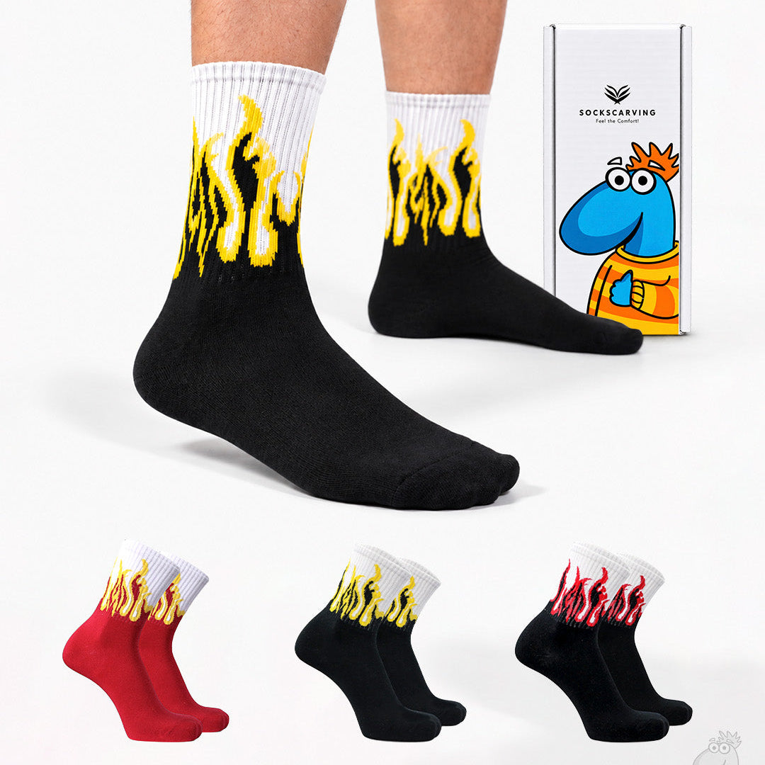 A person wearing black crew socks with a yellow flame design near the top, shown against a light gray studio background. A white product box with a cartoon blue character is visible, along with three smaller color variants at the bottom showing red, black, and black-with-red flame designs.